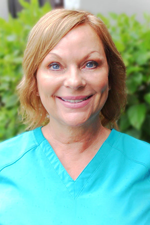 Woman in teal scrubs smiling at camera with green foliage background, dental professional portrait.