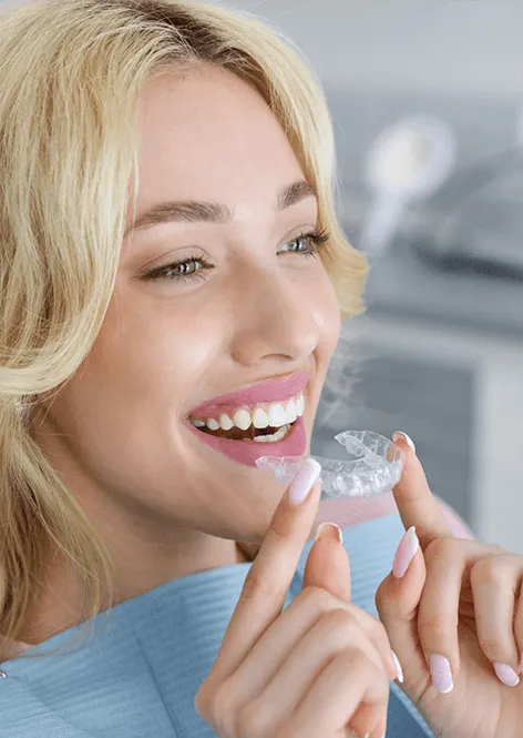 Young woman smiling while holding clear dental aligner in modern clinical setting.