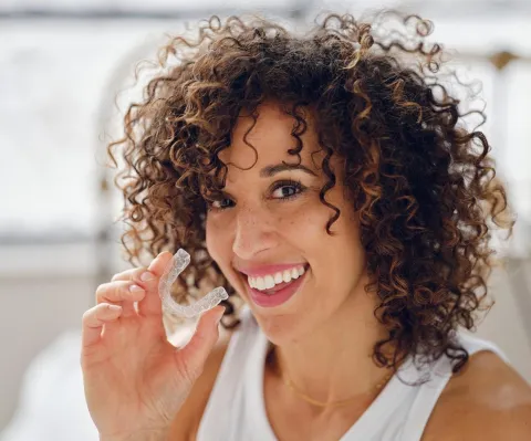 Woman with curly hair smiling while brushing teeth in bright bathroom setting.