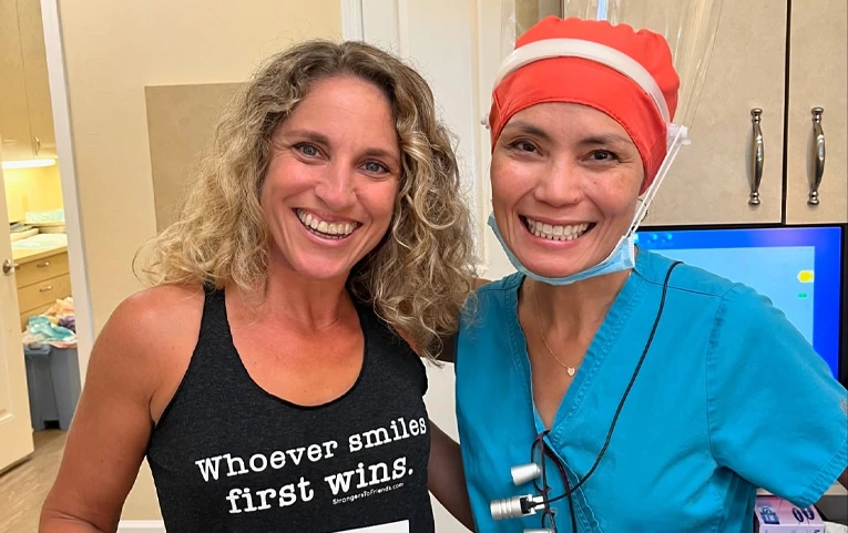 Two women smiling at dental office, one wearing red holiday hat, one in blue scrubs.