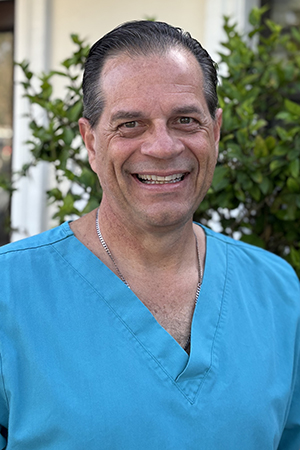 Male dentist in blue scrubs smiling outdoors with greenery background.