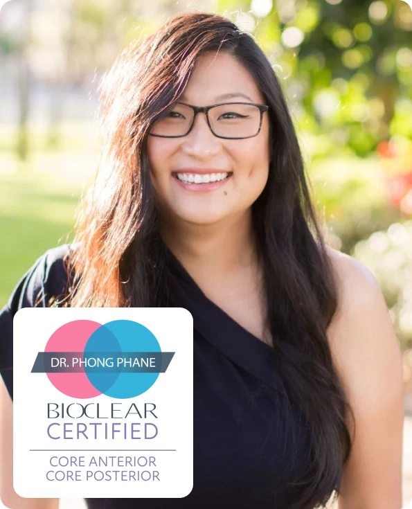 Young woman wearing glasses smiling outdoors, holding Dr. Phong Pham BioClean Certified dental certificate.
