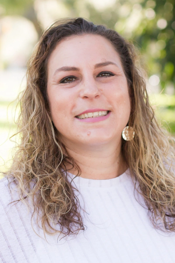 Woman smiling at camera outdoors wearing white shirt with blurred green background.